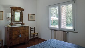 Quarto com cômoda de madeira, uma cadeira, uma cama, janela com vista para vegetação e radiador.