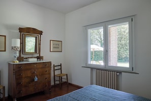 Ein Schlafzimmer mit einem Holzkasten, einem Sessel, einem Bett, einem Fenster mit Blick auf Grünflächen und einer Heizung.