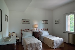 A bedroom with two single beds, a bedside table with a lamp, a small table with a decorative bowl, and framed pictures on the wall.