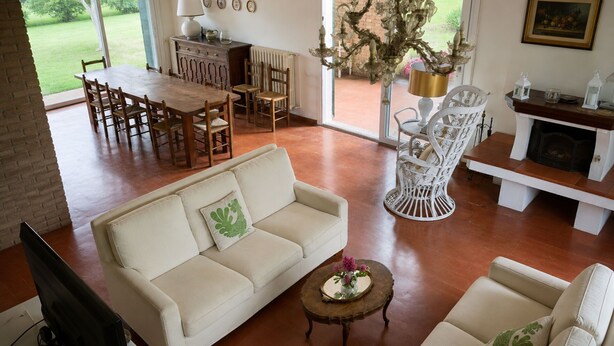 Uma sala de estar espaçosa com um sofá branco, uma mesa de centro e uma poltrona branca. Há uma área de jantar com cadeiras de madeira e uma mesa, e uma lareira com manto branco.
