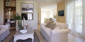 A modern living room with a white sofa, a coffee table with decorative items, a TV, and a bookshelf with various objects. - The Pool Villa In Forte dei Marmi (Forte Dei Marmi)