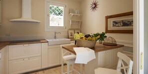 A kitchen with white cabinets, a wooden countertop, a sink, and a window with a lattice design. There is a basket of fruit on the countertop and a framed picture on the wall. - The Pool Villa In Forte dei Marmi (Forte Dei Marmi)