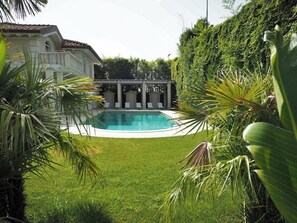 Une piscine entourée d’une végétation luxuriante et un bâtiment blanc orné de colonnes.