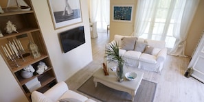 Ein Wohnzimmer mit einem weißen Sofa, einem Couchtisch mit einer Pflanze und einem an der Wand montierten Fernseher.