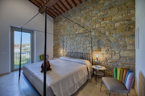 Ein Schlafzimmer mit Steinwand, einem Himmelbett, einem kleinen runden Tisch und einem farbenfrohen Stuhl.