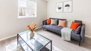 Una sala moderna con un sofá gris, cojines naranja y verde, una mesa de centro de vidrio y dos cuadros abstractos enmarcados en la pared.