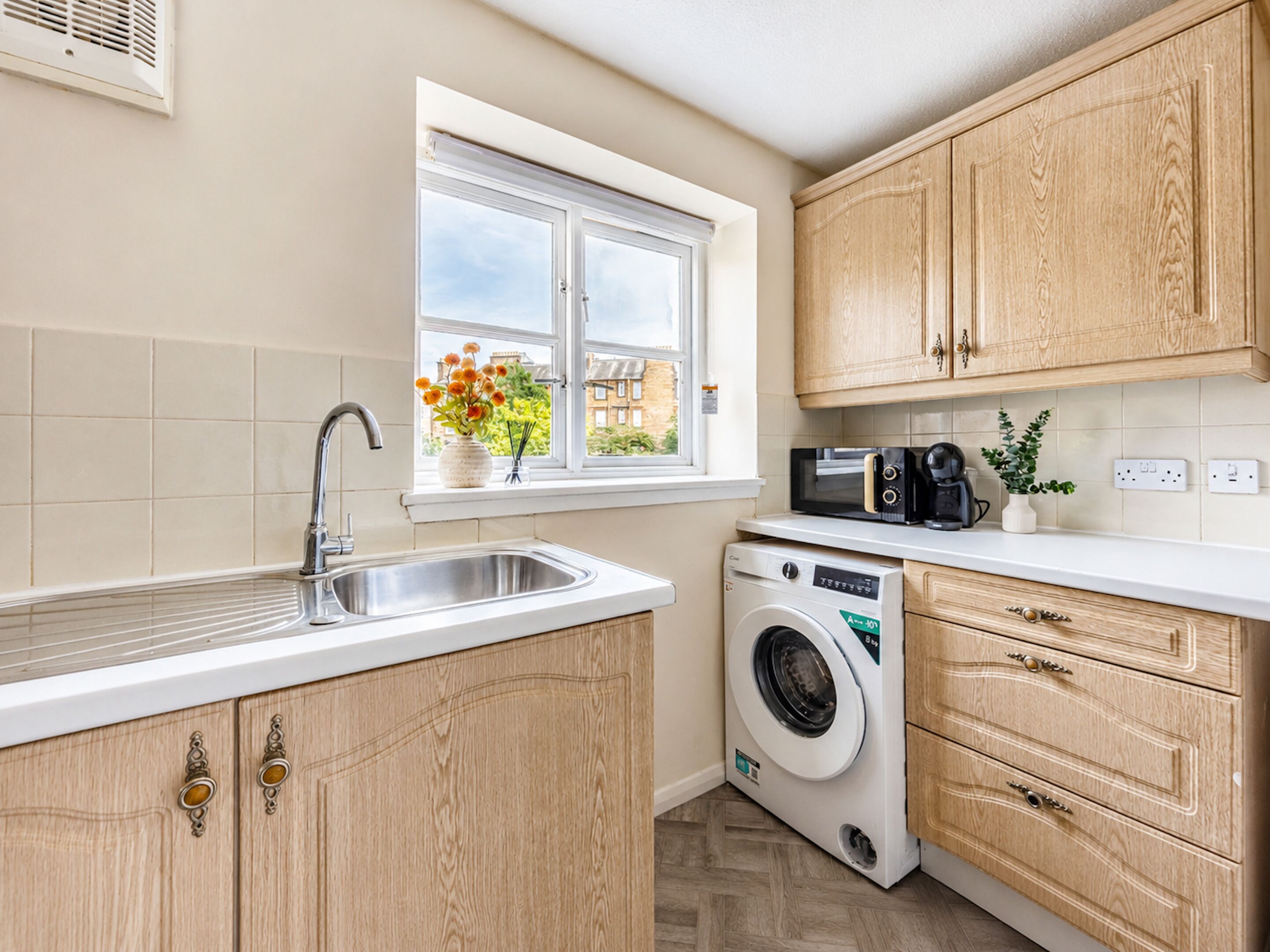 Une cuisine avec des meubles en bois, un lave-linge, un four à micro-ondes et une fenêtre décorée de fleurs.