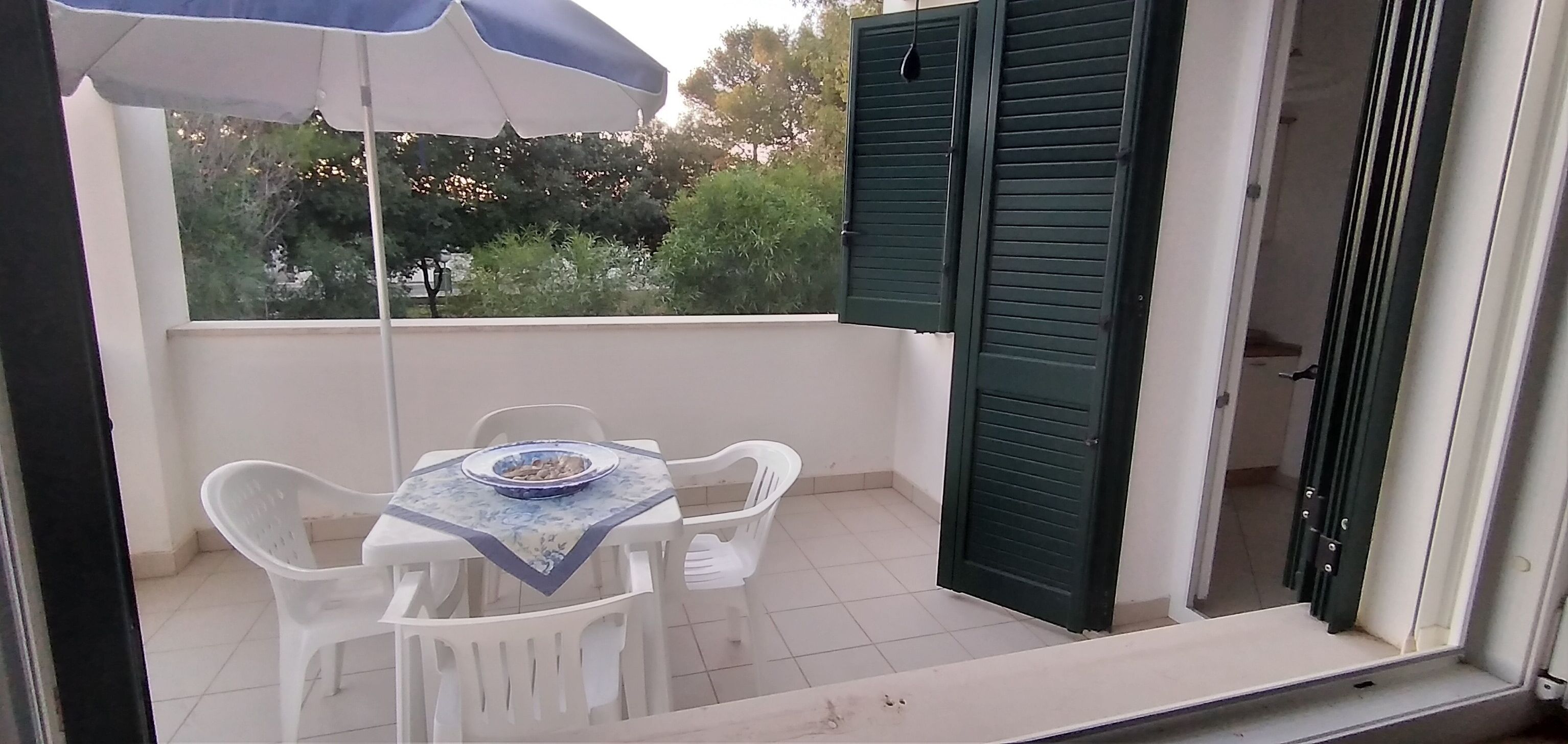 Een balkon met een tafel en stoelen, een blauw-witte parasol en een deur met groene luiken.