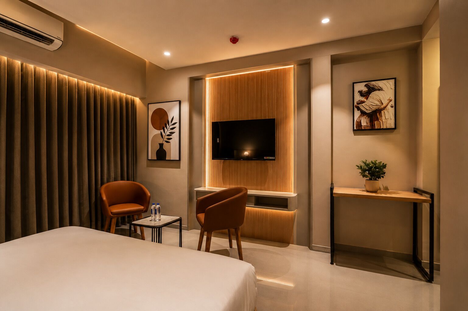 Quarto de hotel com cama, duas poltronas, uma mesinha de centro com garrafas de água, uma TV fixada em um painel de madeira e uma planta em uma mesinha lateral.