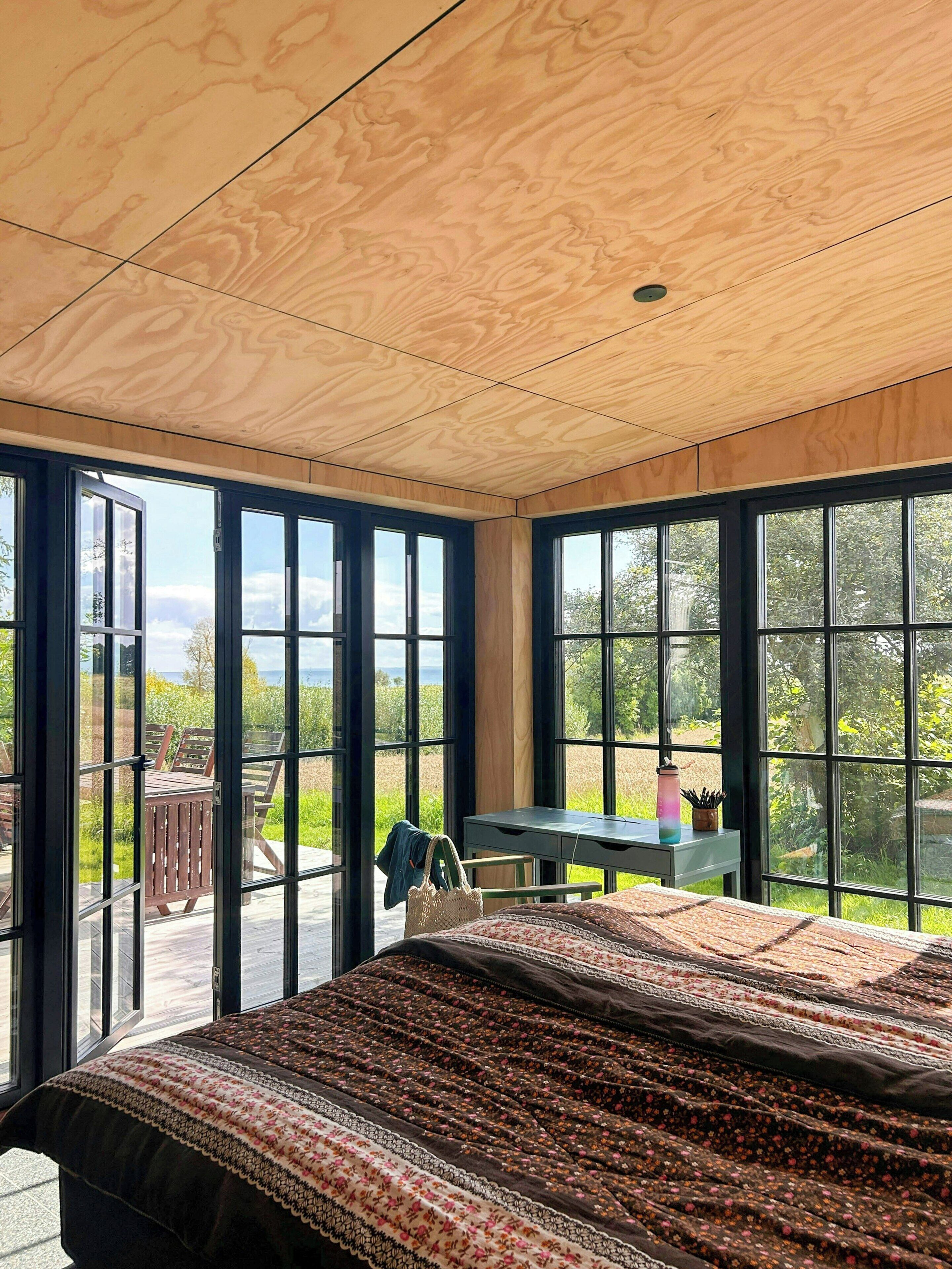 A bedroom with a large bed, wooden ceiling, and large windows offering a view of the outdoors.