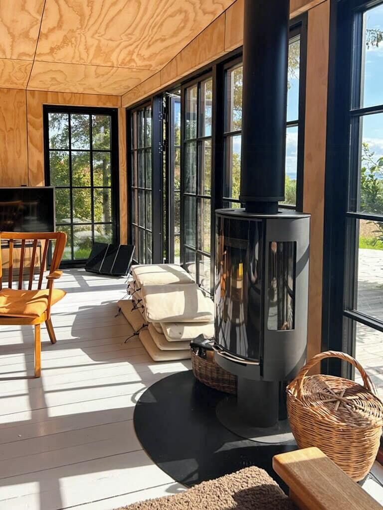 A cozy room with a large window, a black wood-burning stove, a wooden chair, and a small table.