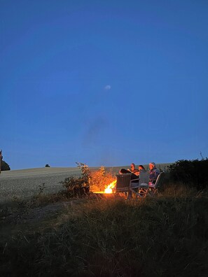 Ein Lagerfeuer mit zwei Personen, die auf Stühlen sitzen, umgeben von Gras und einem Feld.