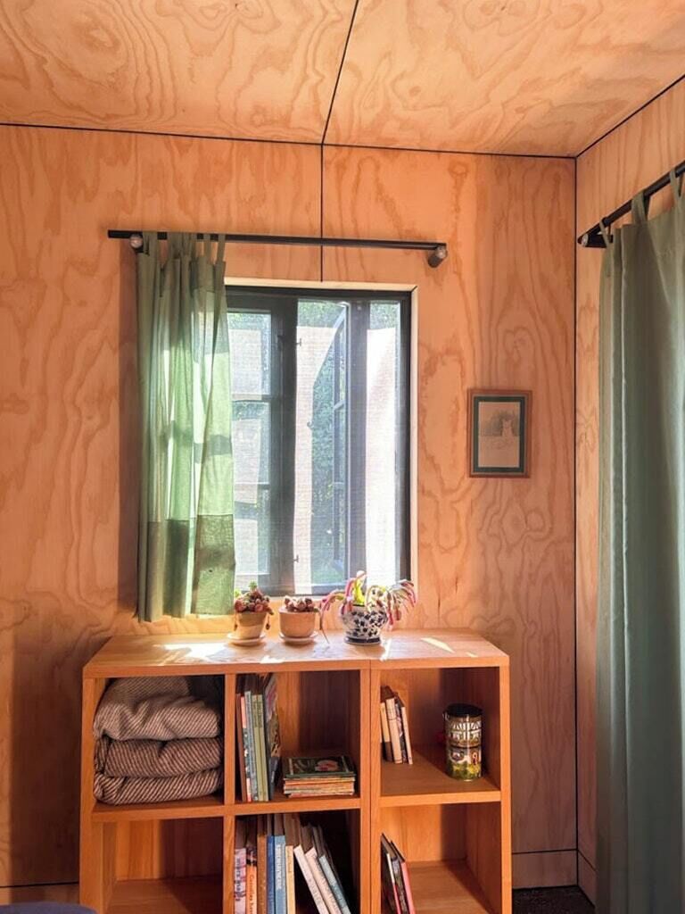 A room with wooden walls, a window with green curtains, a small table with books and decorative items, and a framed picture on the wall.