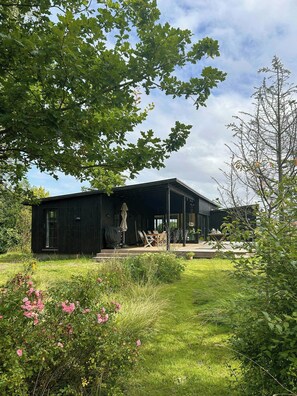 A black wooden building with a covered patio area, surrounded by greenery and flowering plants. - Beach house with sea view in Røsnæs (Kalundborg)
