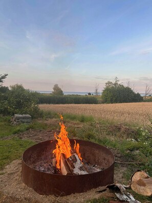 A metal fire pit with flames, surrounded by grass and a field in the background. - Beach house with sea view in Røsnæs (Kalundborg)