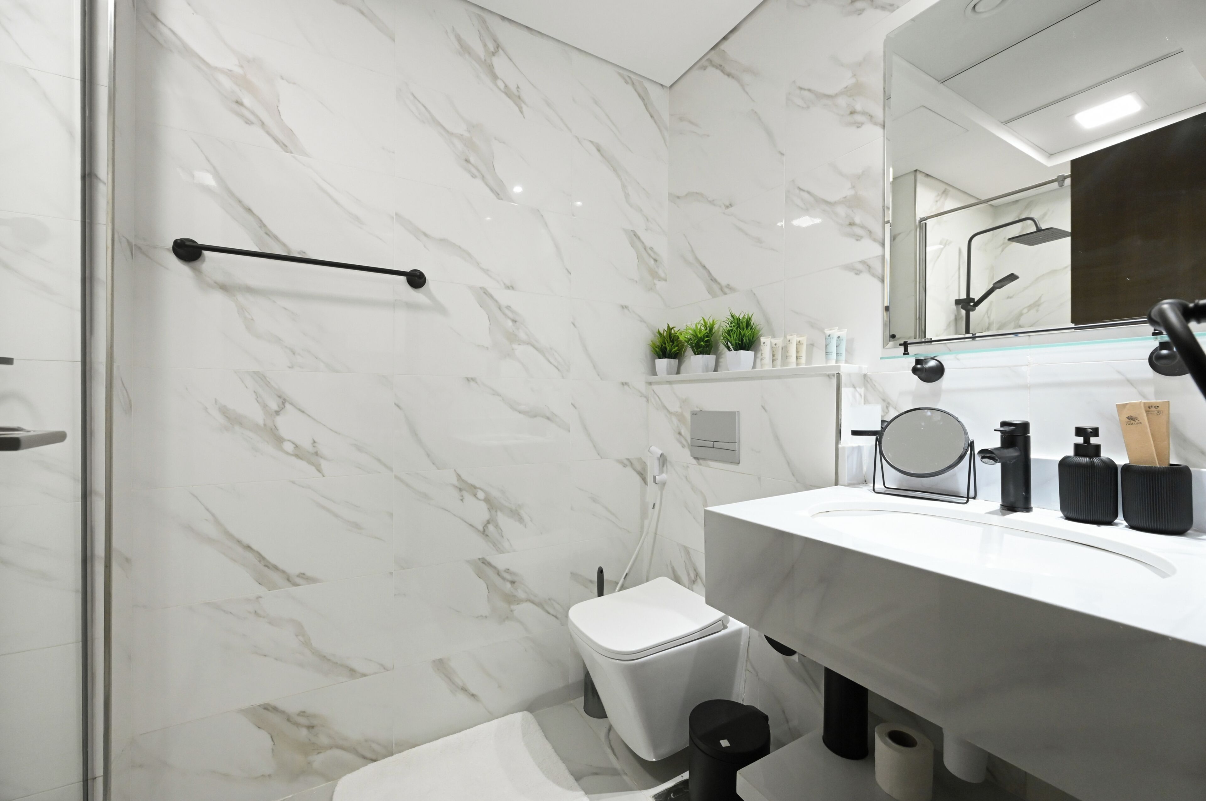 A modern bathroom with a white marble wall, a glass shower enclosure, a white sink with black fixtures, and a toilet.