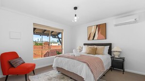 Un dormitorio con cama, una silla, una mesita de noche, lámpara, un cuadro y una ventana con vistas a edificios.