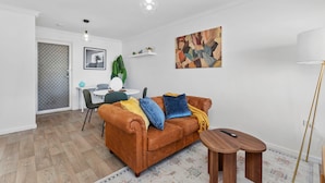 Una sala de estar con un sofá marrón, una mesa de centro, una lámpara, un cuadro y una zona de comedor con una mesa y sillas.