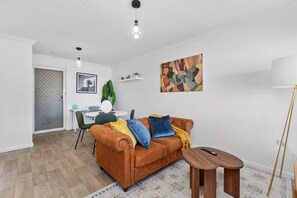 A living room with a brown sofa, a coffee table, a lamp, a painting, and a dining area with a table and chairs. - Dashing Dome - A Sepal Stay (Perth)