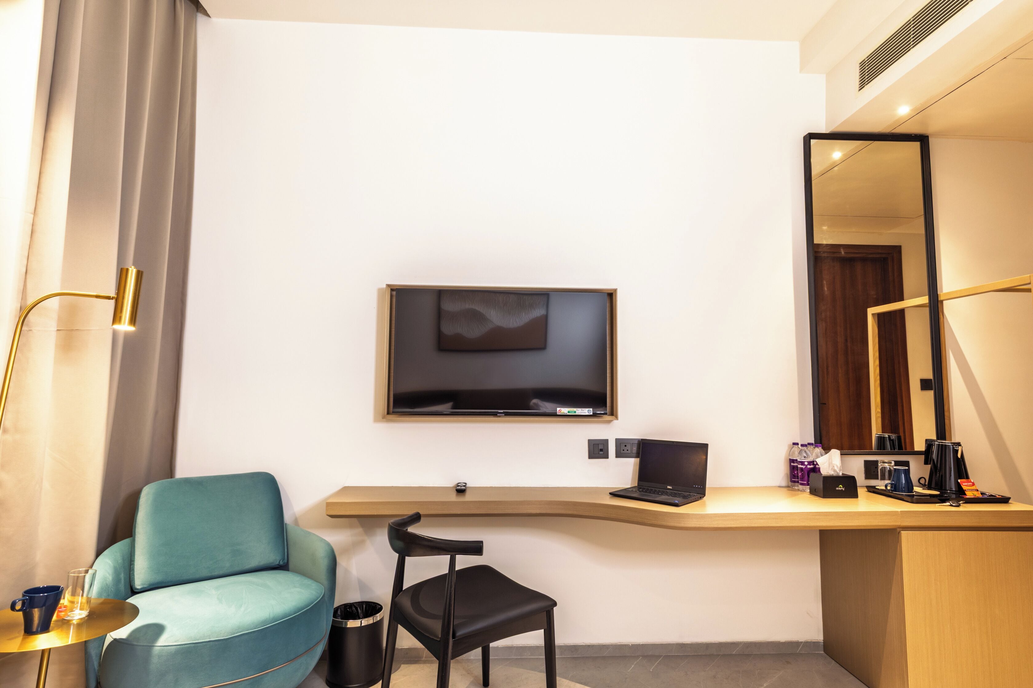 Habitación de hotel con televisión de pantalla plana montada en la pared, escritorio con computadora portátil, silla, cesto de basura y cafetera.