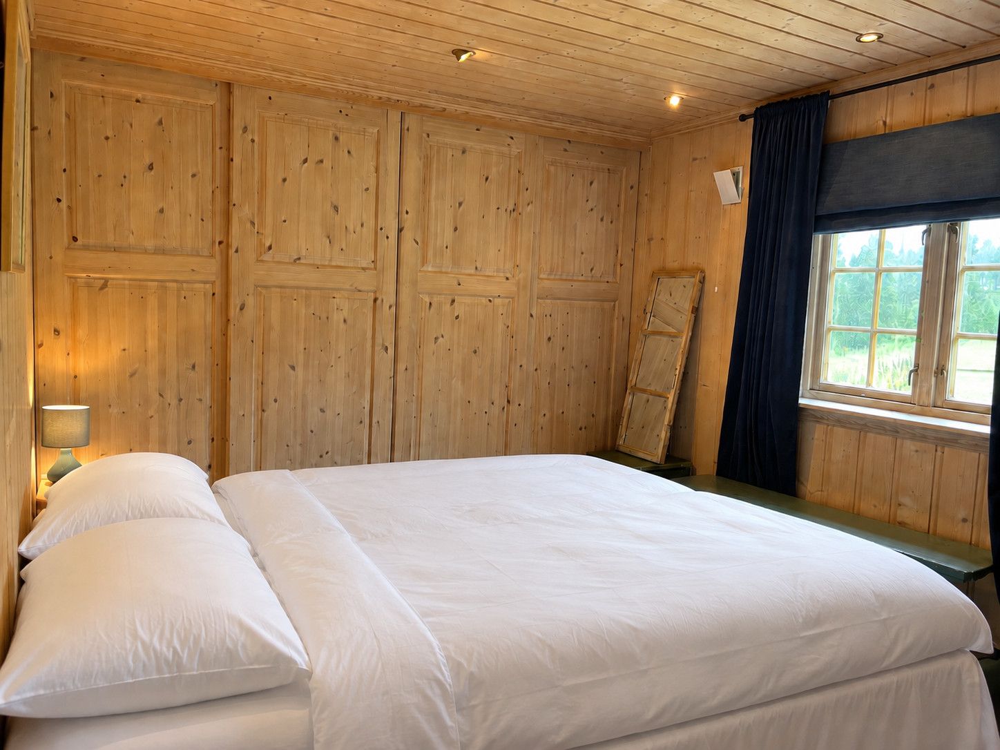 Habitación con paneles de madera, una cama, una mesita de noche y un ventanal con cortinas.