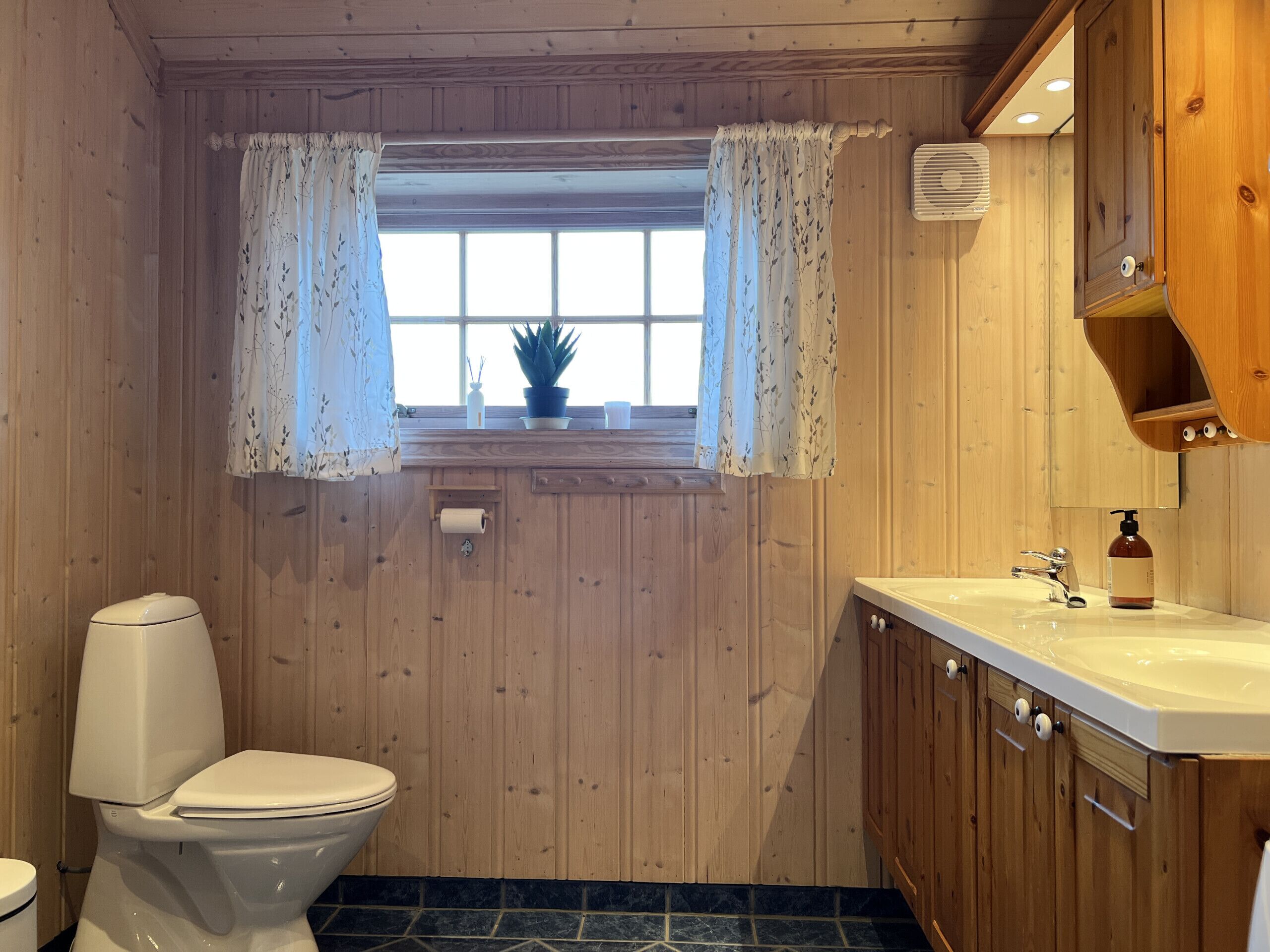 A wooden-paneled bathroom with a toilet, a window with curtains, a sink with a mirror, and a small shelf with toiletries.