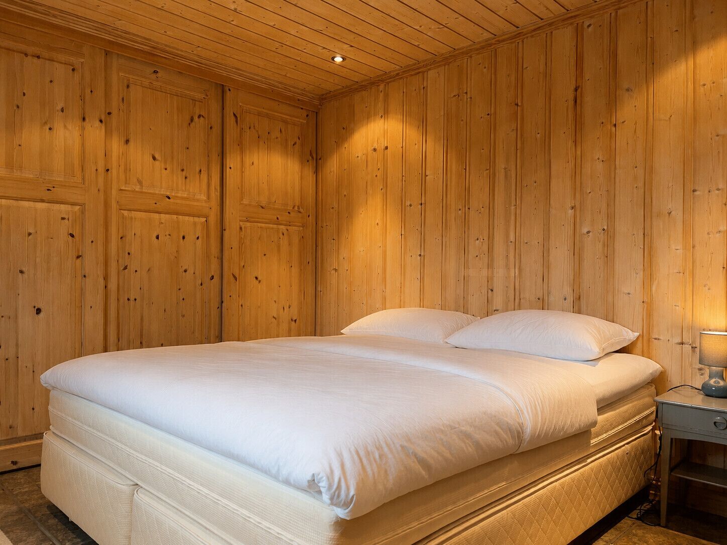 Une chambre avec un lit simple, des murs recouverts de panneaux de bois, une table de chevet avec une lampe et un matelas avec du linge de lit blanc.
