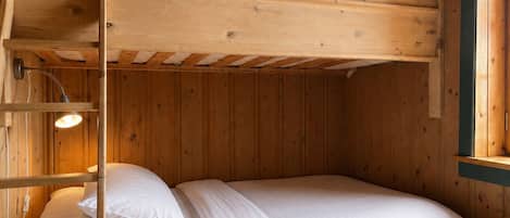 Un lit superposé avec une tête de lit en bois et un lit simple en dessous. Une lampe fixée au mur se trouve sur le côté gauche.