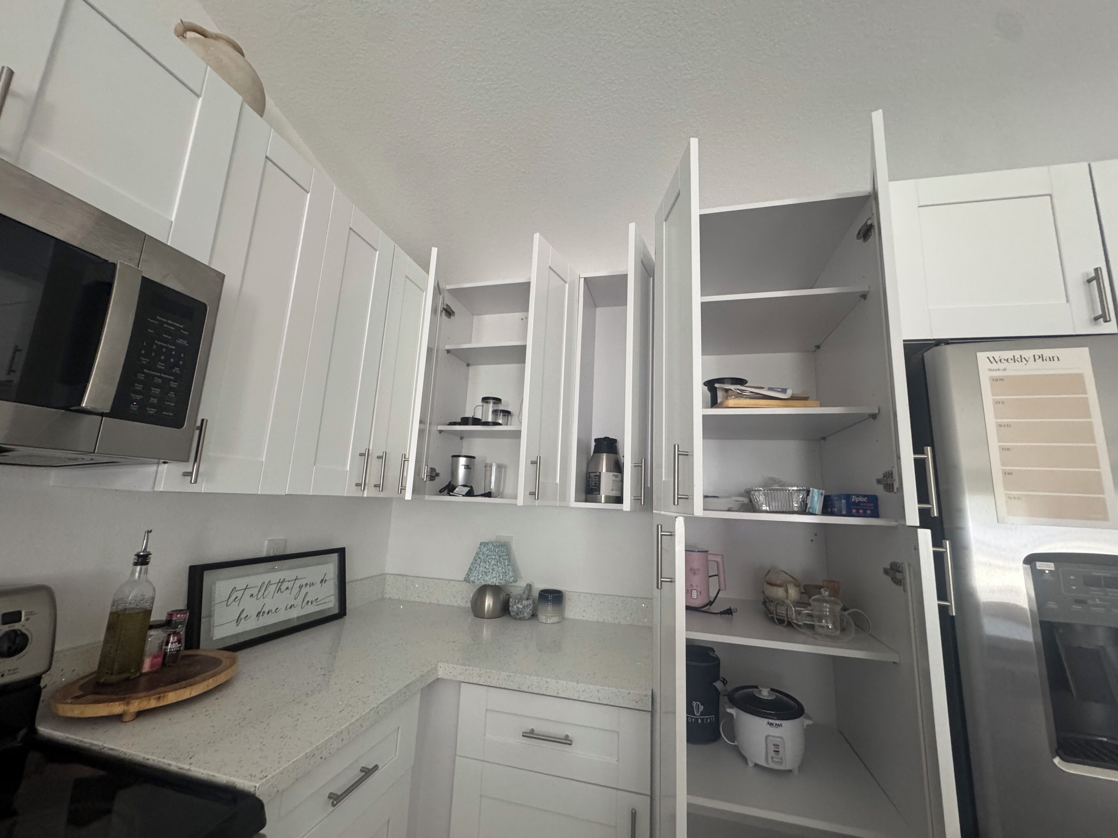 A kitchen with white cabinets, a microwave, a refrigerator, and a weekly planner on the door.