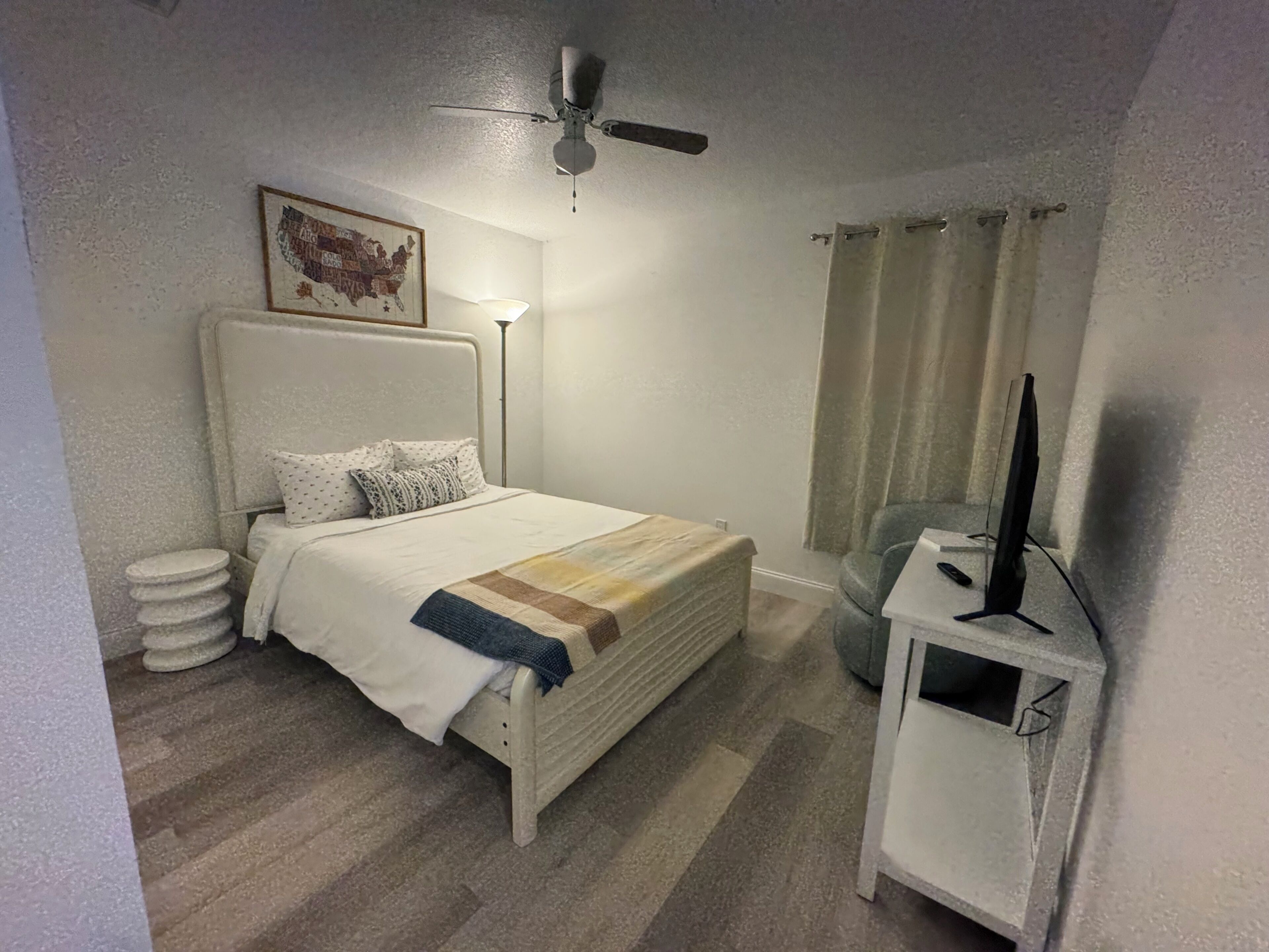 Une chambre à coucher avec un lit, une lampe de chevet, un ventilateur de plafond, un téléviseur posé sur un meuble et une carte accrochée au mur.