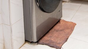 En kompakt tvättmaskin i rostfritt stål med frontmatning och en digital kontrollpanel, placerad i ett kök med vita kakelplattor på golvet och en brun matta.