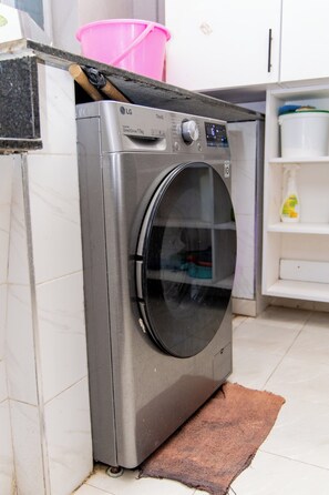 Eine kompakte, frontgeladene Waschmaschine aus Edelstahl mit digitaler Bedienblende, platziert in einer Küche mit weißen Fliesen und einem braunen Teppich.