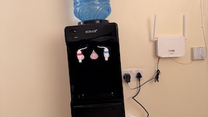 Ein SONAR-Wasserspender mit einer großen Wasserflasche obenauf, eine Steckdose und ein an der Wand befestigtes Gerät.