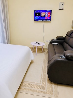 A hotel room with a bed, a sofa, a small table, and a wall-mounted TV displaying streaming services. - Cozy Homes Airbn. 15 minutes from JKIA in Kasarani, Nairobi (Nairobi)