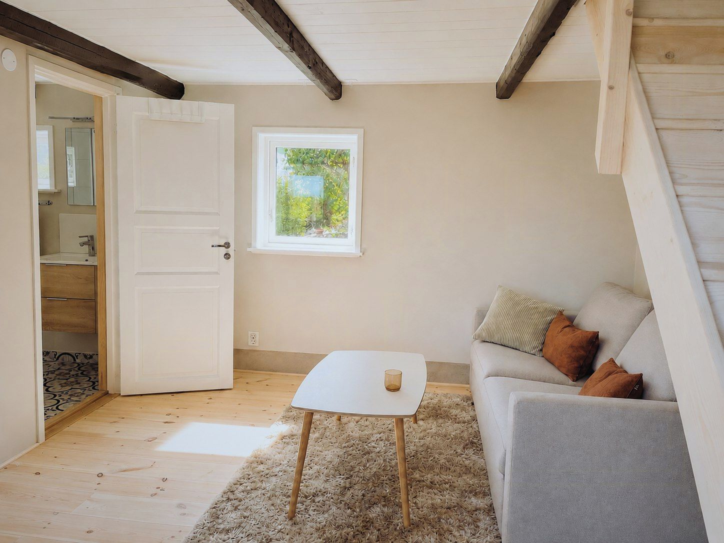 A room with a white door, a small table, a grey sofa with cushions, a window with a view of greenery, and a wooden floor.