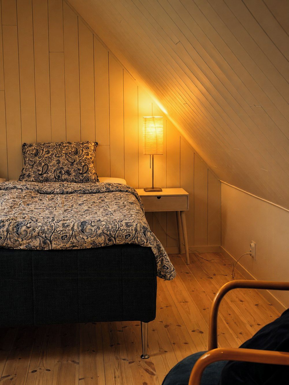 A bedroom with a bed, bedside table, lamp, and a chair.