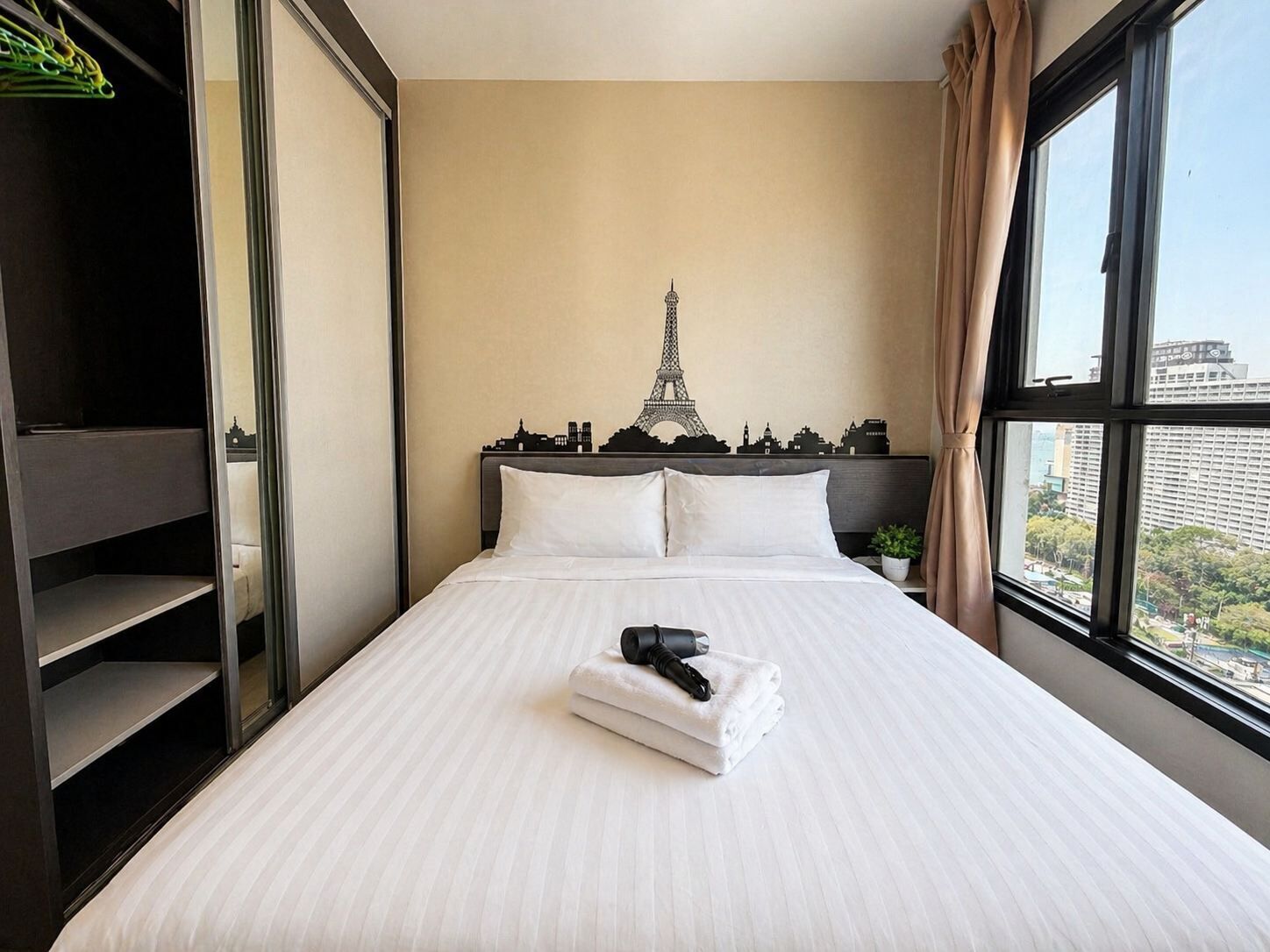 A hotel room with a bed, a view of the Eiffel Tower silhouette on the wall, a window with a cityscape view, and a shelf with a plant.