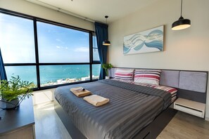 A modern bedroom with a large window, a bed with striped bedding, a headboard, and a wall-mounted abstract painting. - WCC The Base Central Pattaya (Bang Lamung)