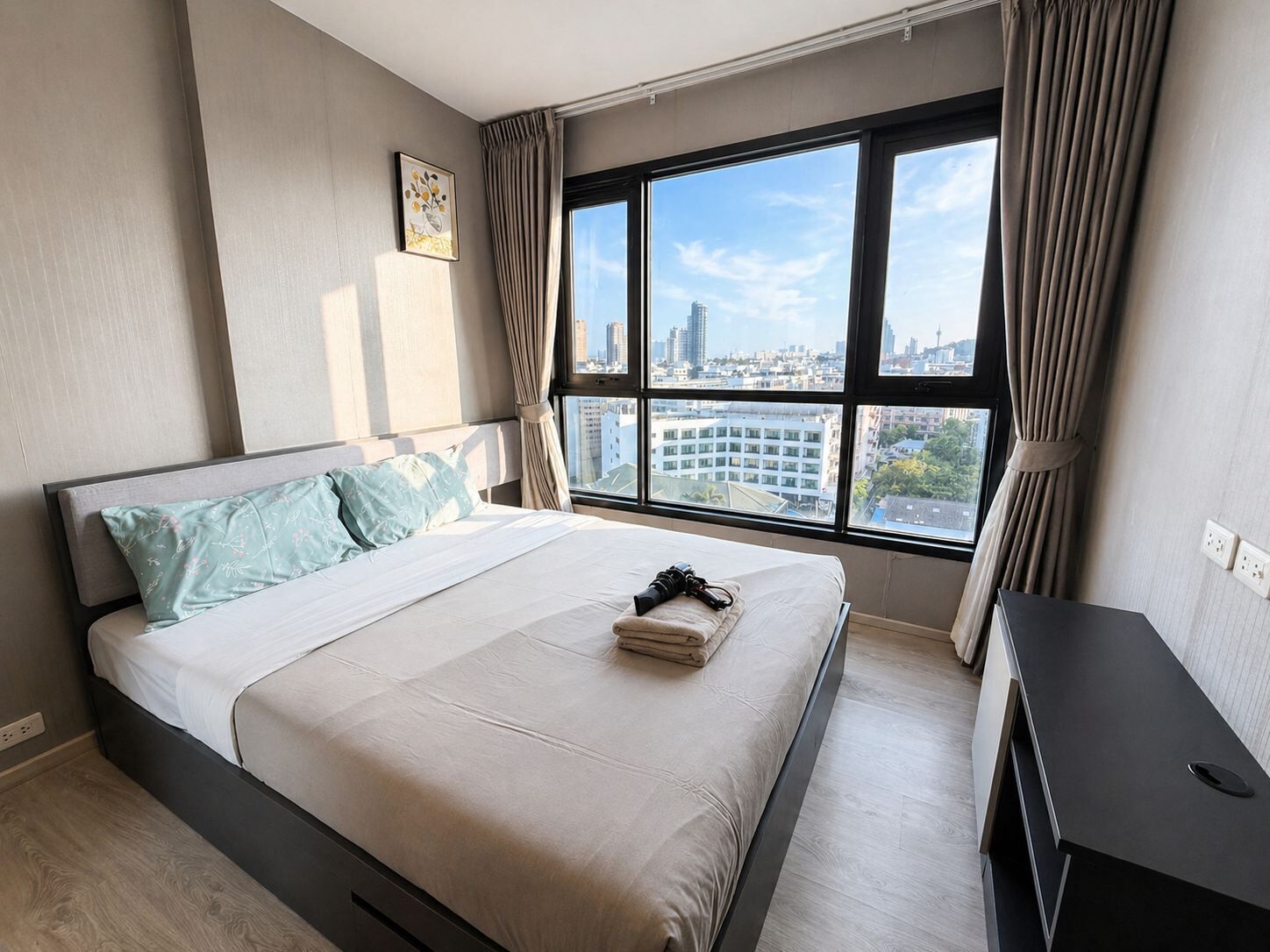 A modern hotel room with a large window, a bed with a white and light blue bedspread, a small bedside table, and a city view.