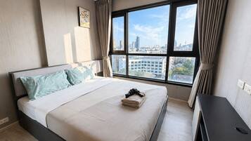 A modern hotel room with a large window, a bed with a white and light blue bedspread, a small bedside table, and a city view.