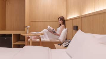 A person sitting on a bed in a room with wooden walls, reading a book. There is a bedside table with a lamp and a phone.