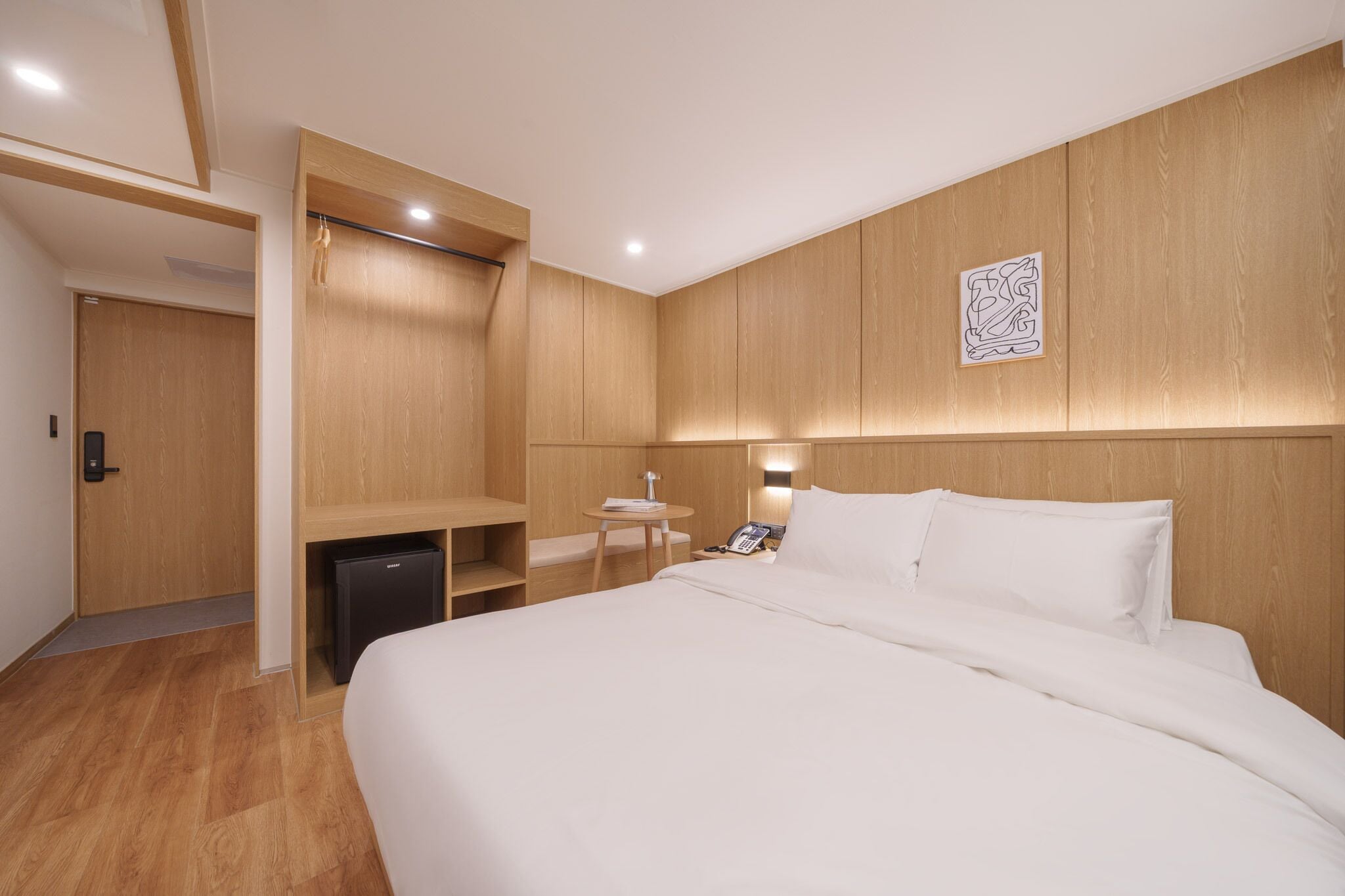 A modern hotel room with a large bed, a desk, a chair, and a small table with a lamp.