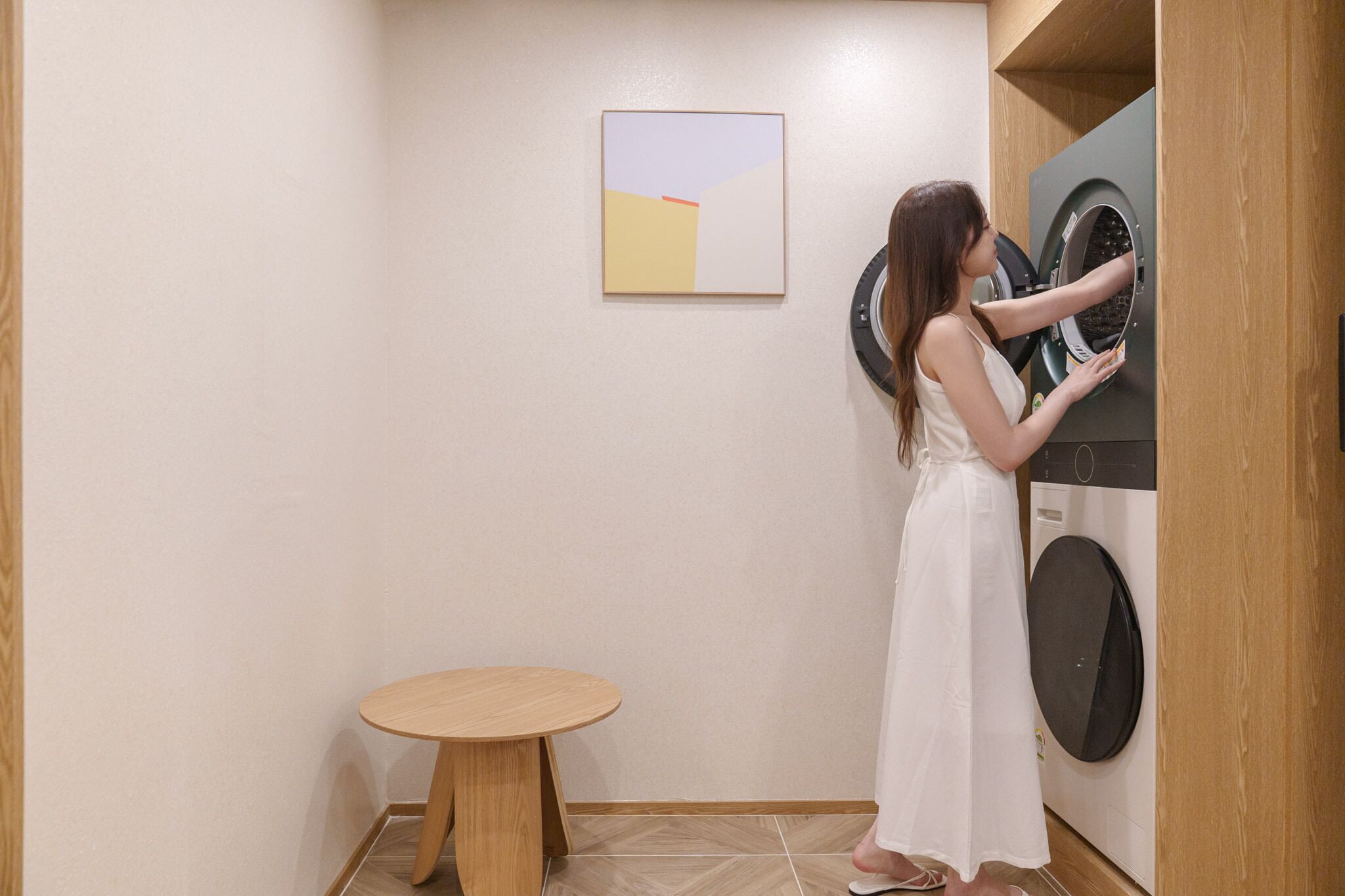 A person in a white dress standing next to a washing machine in a room with a wooden table and a painting on the wall.