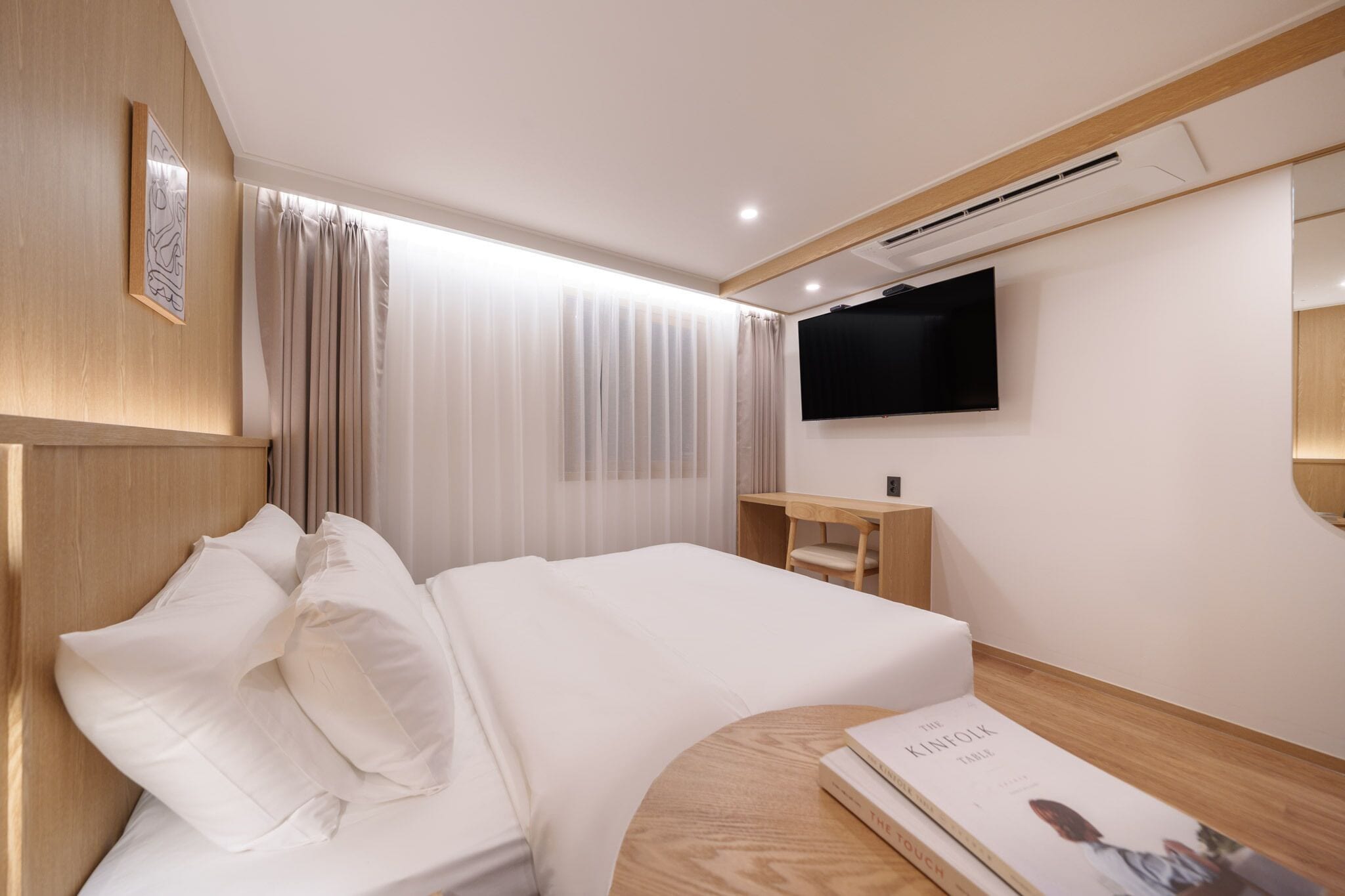 Una habitación de hotel moderna con una cama grande, una mesita de noche, un televisor de pantalla plana montado en la pared y un amplio ventanal con cortinas traslúcidas.