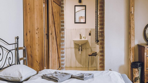Une chambre avec une armoire en bois, un lit avec une literie blanche, une petite salle de bain avec un lavabo et un miroir, et une arche en briques.