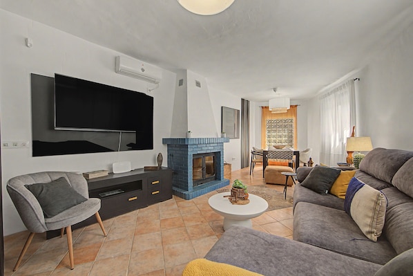 A modern living room with a flat-screen TV, a blue fireplace, a grey sofa, and a dining area with a table and chairs.