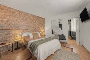 A bedroom with a stone wall, a bed with a blanket, a bedside table with a lamp, a chair, and a TV mounted on the wall.