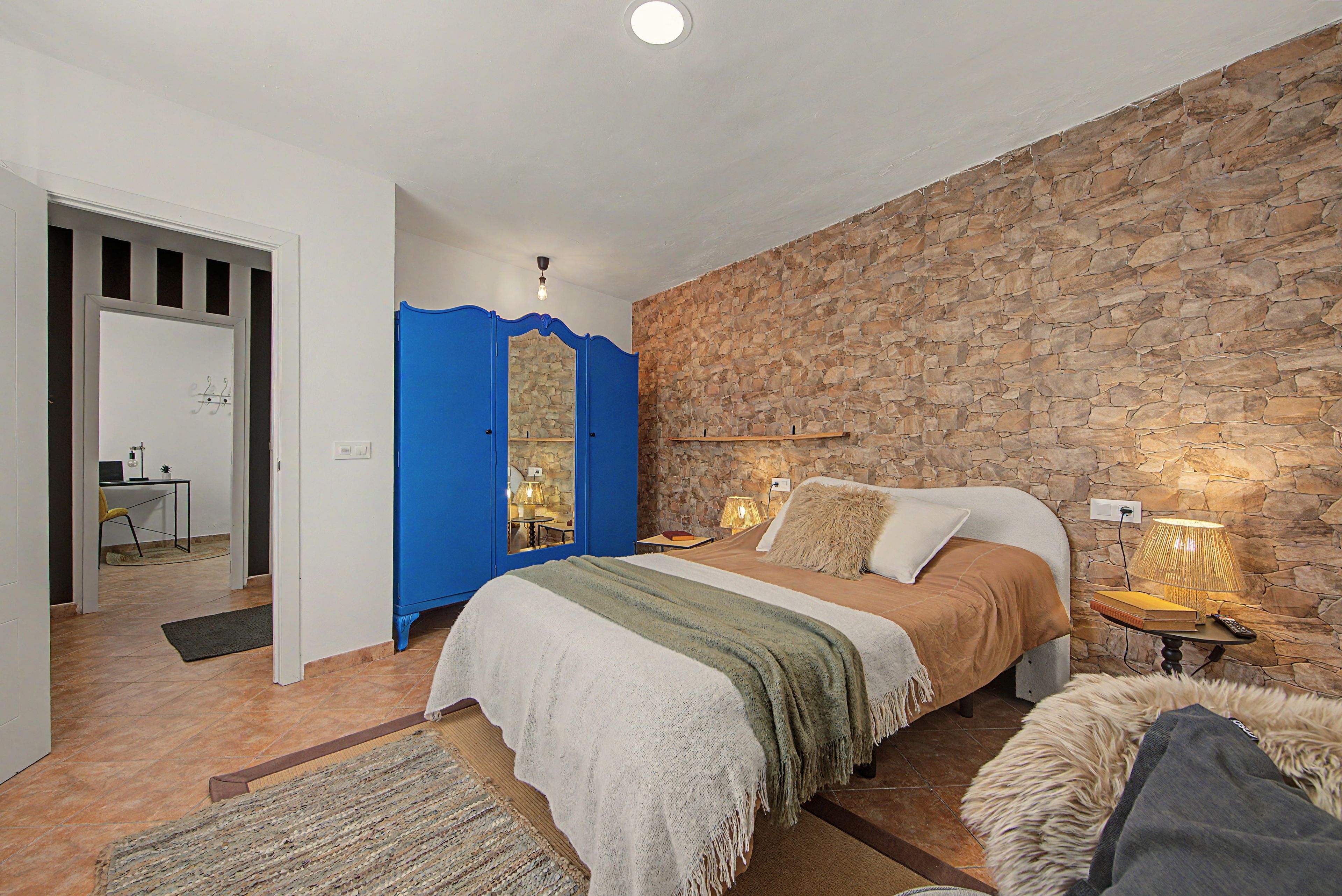 A bedroom with a stone wall, a bed with a green blanket, a desk with a lamp, and a chair.