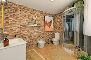 A bathroom with a stone wall, a shower with a glass door, a toilet, a sink, and a small shelf with plants.