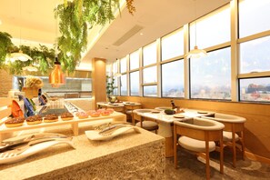 A modern restaurant interior with a buffet counter, seating area, and large windows offering a city view.